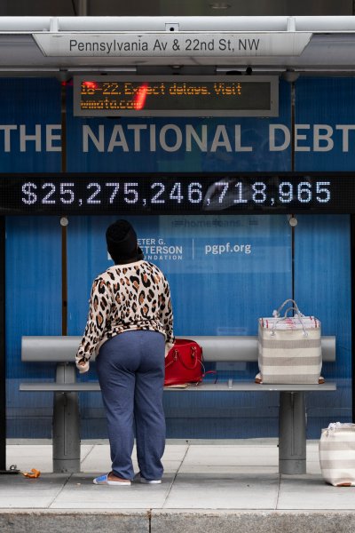 Passengers wearing face masks wait for their bus in front of a national debt display on Pennsylvania Ave. NW in Washington.