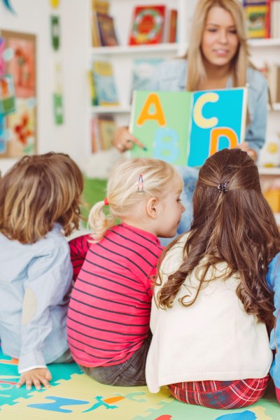 Teacher with a group of preschool children in a nursery. The children are sitting on the floor and listening teacher. Learning letters. In the background we can see a shelf with some, toys, black board and books. View from behind.