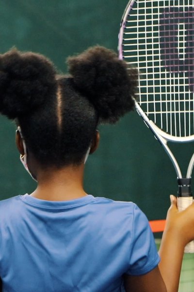 Young Black girl with buns holds up a tennis racket.