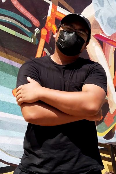 Masked man stands with arms folded in front of mural of bicycle.