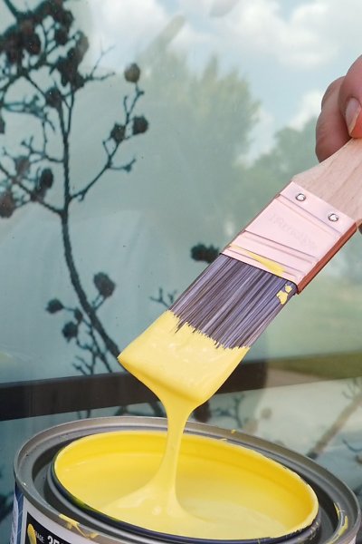 Paint brush drizzles yellow paint back into a can in front of a window.