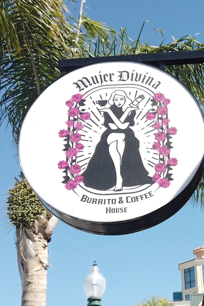 A sign hangs in front of a shop reading "Mujer Divina Burrito and Coffee House" with illustration of woman holding coffee and a burrito.