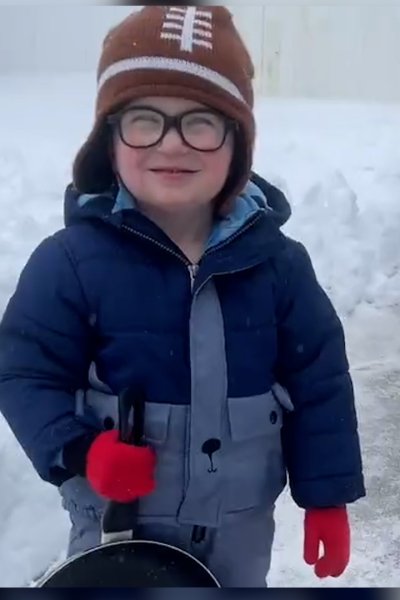 Little boy smiles in snow gear holding frying pan.