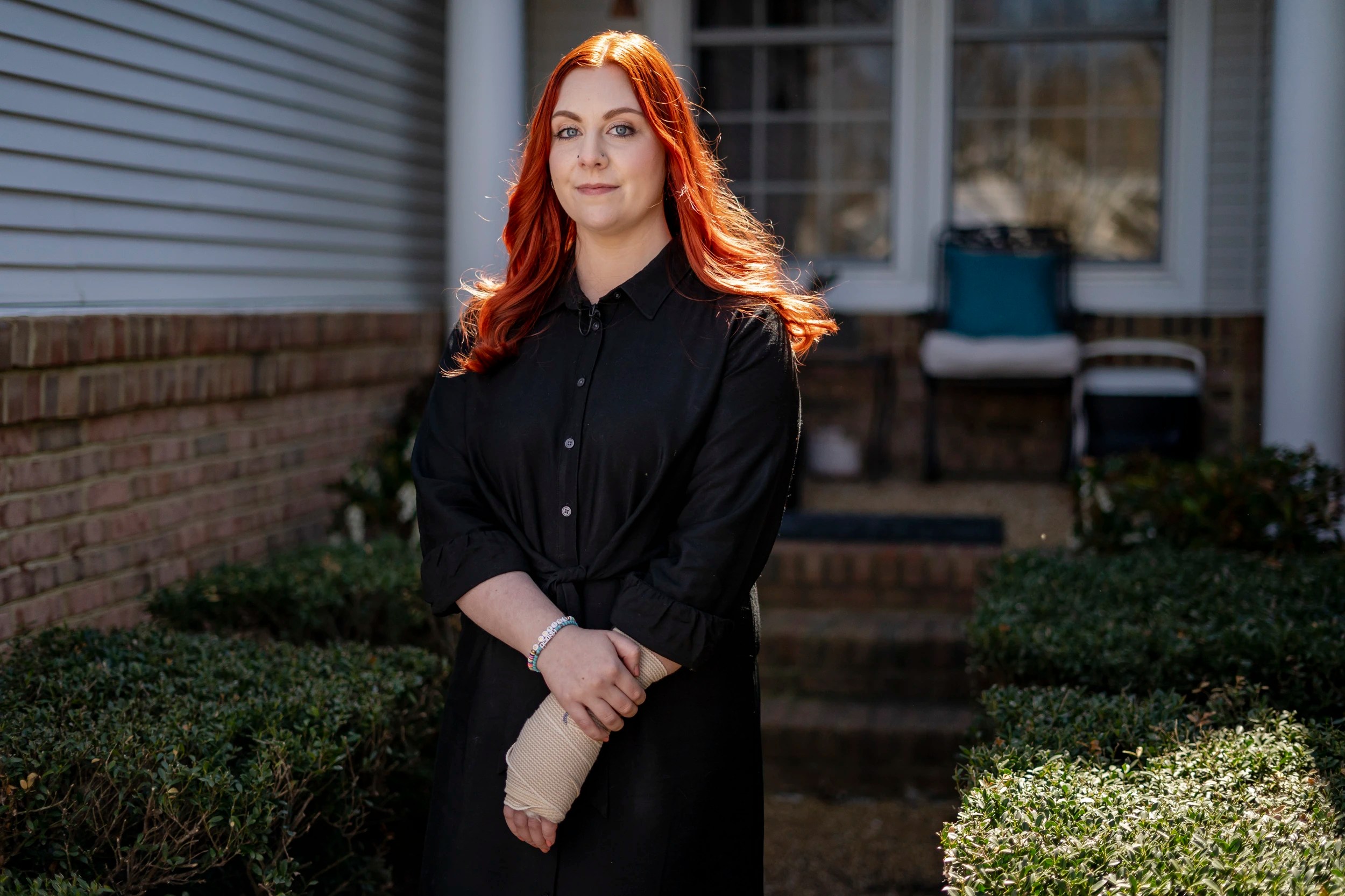 A photo of Abigail Zwerner. She has red hair and is wearing a black blouse, standing against the sun in front of a white house. 